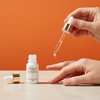 Hand holding a dropper with ARAGAN Secret nail treatment over a white bottle on an orange background